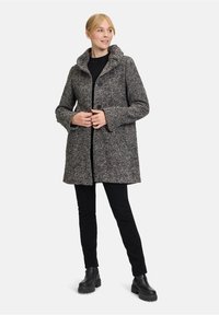 Grijze jas met textuur, hoge kraag, twee knopen en zijzakken, gecombineerd met zwarte broek en stevige zwarte laarzen.