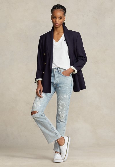 Kvinna som bär en marinblå blazer över en vit V-ringad tröja, ljusblå slitna jeans med rivna knän och vita sneakers, stående mot en neutral bakgrund.