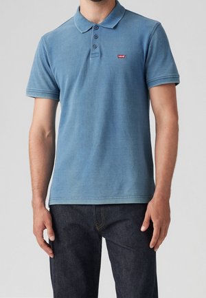 Homme portant un polo à manches courtes bleu avec un logo Levi's rouge, associé à un jean en denim foncé, sur un fond clair uni.