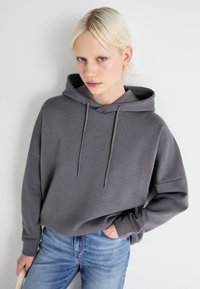 Grå hettegenser med snøring og avslappet passform, kombinert med lyseblå jeans. Glatt materiale og subtile sømdetaljer.