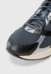 Moška tekaška obutev z zračnim sivim mrežastim zgornjim delom, črnimi usnjenimi akcenti, srebrnimi detajli in teksturiranim gumijastim podplatom.