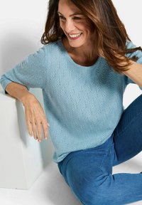 Hellblauer Strickpullover mit einem strukturierten Muster und Dreiviertelärmeln, kombiniert mit blauen Jeans, vor einem weißen Hintergrund sitzend.