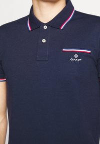 Granatowa koszulka polo z bawełny, z kołnierzykiem w kontrastowe paski, zapięciem na trzy guziki oraz wyszywanym logo na kieszeni na piersi.
