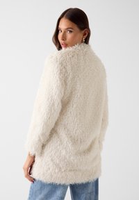 Wollige, crèmekleurige jas met een gestructureerd, zacht oppervlak, lange mouwen en een ontspannen pasvorm, die tot onder de heupen reikt.