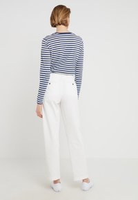 Person med kort hår iklädd en blå och vit randig långärmad skjorta, vita högmidjade byxor och vita sneakers, vänd bort från kameran mot en vit bakgrund.