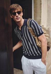 Marinrandig polo, kort ärm, och ribbad krage. Vita linnebyxor och en brun tygryggsäck. Solglasögon med bruna ramar.