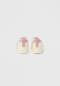 White sneakers with light pink suede heel tabs, textured rubber soles, and minimalistic design. Smooth material with a rounded shape.