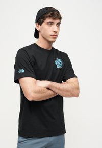 Νέος άνδρας που φορά μαύρο καπέλο με το γείσο προς τα πίσω, μαύρο μπλουζάκι The North Face με μπλε λογότυπο, με σταυρωμένα τα χέρια, κοιτάζει προς το πλάι σε απλό λευκό φόντο.