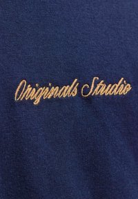 Tkanina tamnoplave boje s mekim teksturom, s izvezenim natpisom "Originals Studio" u zlatnom kurzivu.