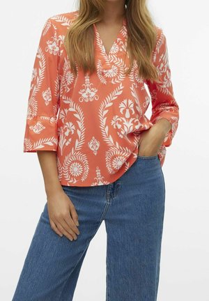 Frau trägt eine orangefarbene Bluse mit V-Ausschnitt, weißen Blumenmustern und blauen Jeans, eine Hand in der Tasche.