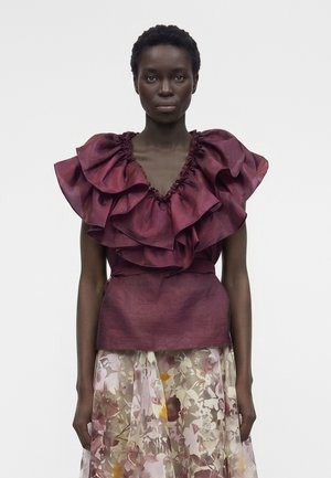 Kobieta w bezrękawniku w głębokim bordowym kolorze z falbankami, zestawionym z przejrzystą kwiatową spódnicą na jednolitym jasnym tle.