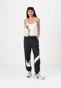 Melna sporta bikses ar baltu diagonālo svītru, pelēks topiņš un bēša plecu soma. Apavi ir bēši un pelēki Nike sporta apavi.