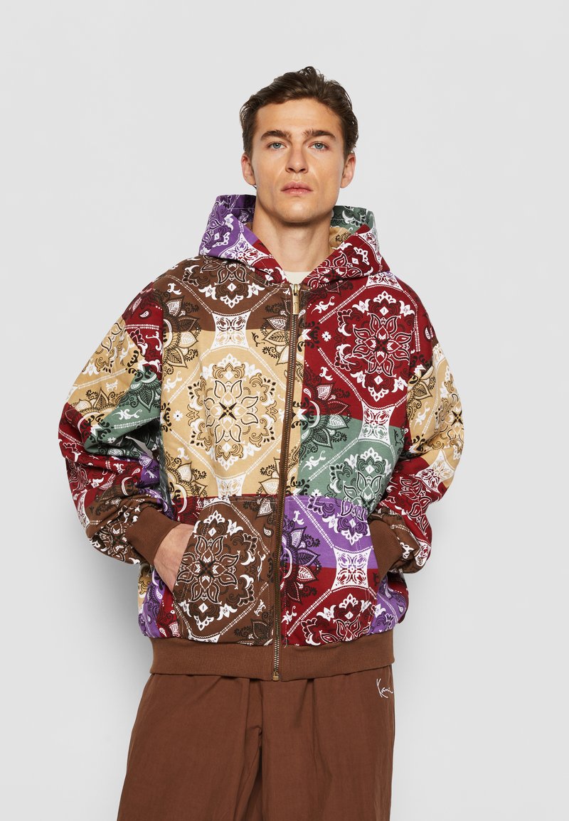 Hooded zip-up jacket featuring a patchwork design with intricate patterns in red, brown, green, purple, and gold tones. Brown ribbed cuffs.