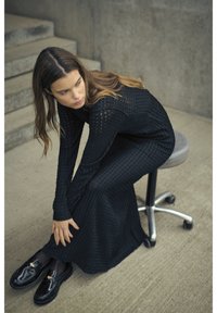 Schwarzes Strickkleid mit strukturiertem Muster, langen Ärmeln und lässigem Schnitt. Schwarze Lackslipper, auf einem Hocker vor einem grauen Hintergrund sitzend.