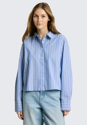 Młoda kobieta ubrana w luźną, niebiesko-białą koszulę w pionowe paski na guziki oraz jasnoniebieskie dżinsy, stojąca na tle jednolitego tła.