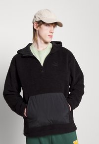 Pullover in pile nero con collo a zip, dotato di tasca sul petto nera trapuntata e cappuccio testurizzato. Indossato sopra una camicia verde chiaro.