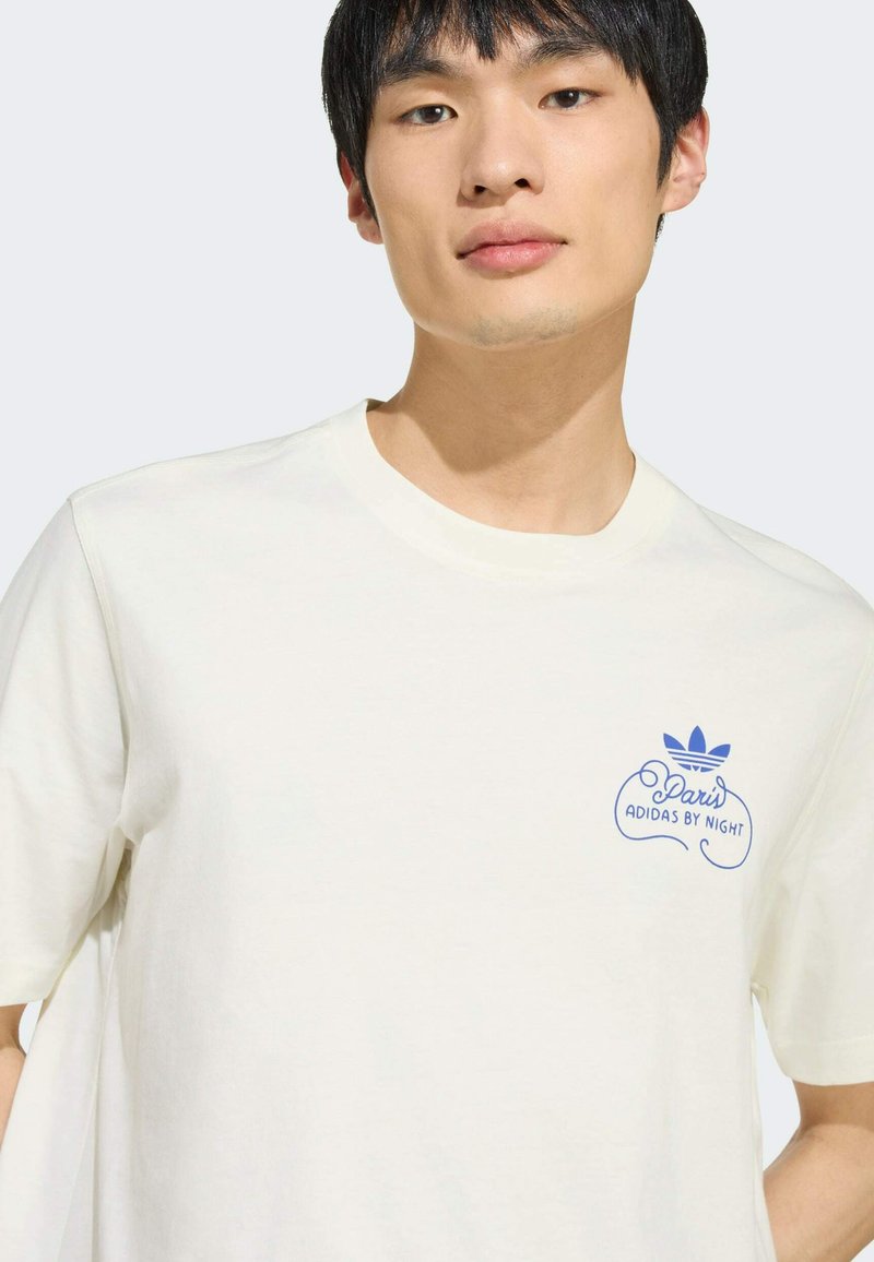 Jeune homme portant un t-shirt écru avec un logo Adidas bleu et le texte "Paris Adidas By Night" sur le côté gauche de la poitrine.