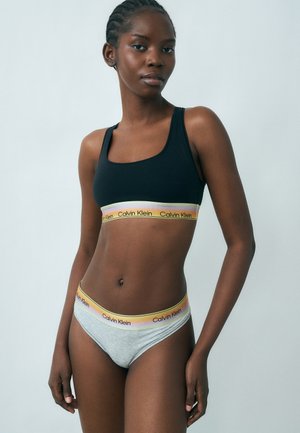 Mladá žena stojaca, oblečená v čiernej športovej podprsenke a sivých nohavičkách s pásmi v dúhových farbách značky Calvin Klein.