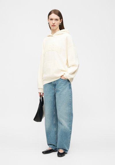 Femme portant un sweat à capuche crème oversize, un jean bleu ample, des ballerines noires, tenant un sac à main noir, debout devant un fond clair uni.