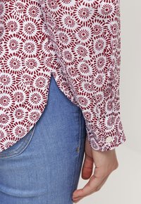 Chemise à manches longues avec un motif floral rouge sur un tissu blanc. La chemise présente un ourlet courbé et un détail bouton au poignet, portée avec un jean bleu.