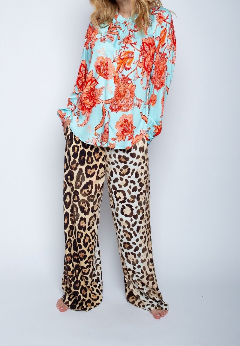 Femme portant une chemise bleue à boutons avec un imprimé floral rouge et un pantalon large à motifs léopard mélangés, debout les mains dans les poches.