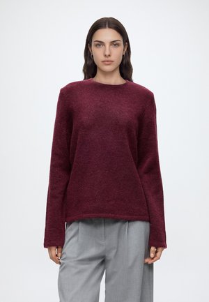Femme aux longs cheveux foncés portant un pull bordeaux duveteux et un pantalon gris clair, debout devant un fond clair uni.
