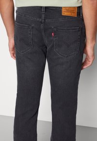 Hombre con vaqueros negros de denim con dos bolsillos traseros, etiqueta de marca roja y parche de piel marrón en la cintura.