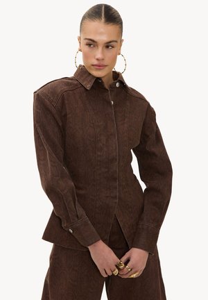 Femme aux cheveux plaqués en arrière, portant une veste boutonnée marron texturée et un pantalon assorti, accessoirisée de grandes boucles d'oreilles en anneau et de bagues en or.