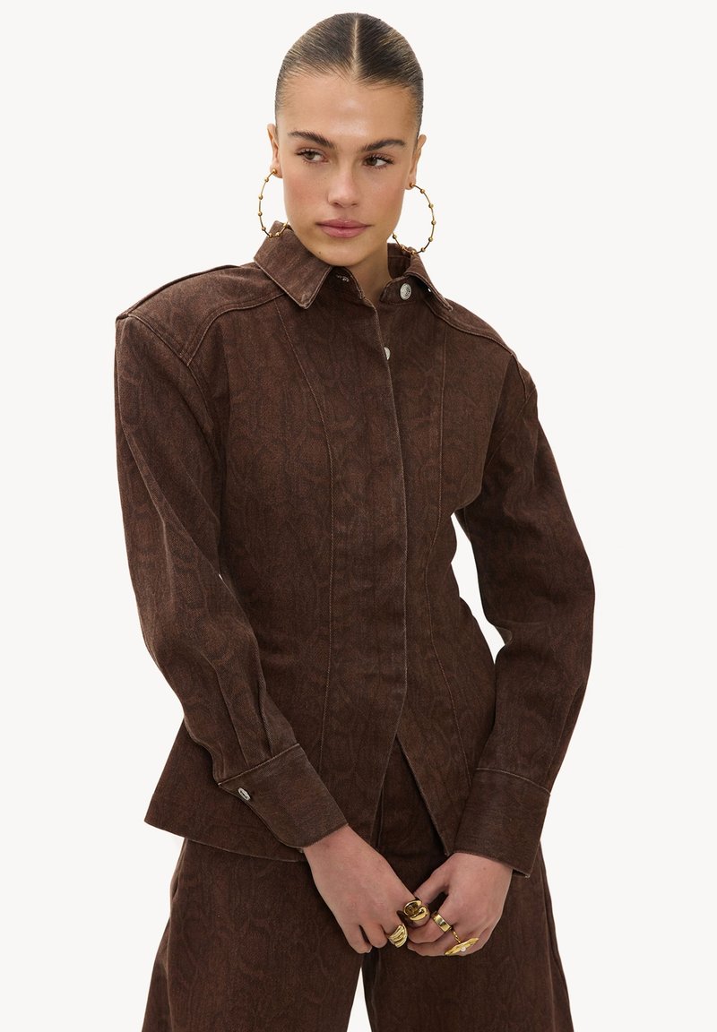 Femme aux cheveux plaqués en arrière, portant une veste boutonnée marron texturée et un pantalon assorti, accessoirisée de grandes boucles d'oreilles en anneau et de bagues en or.