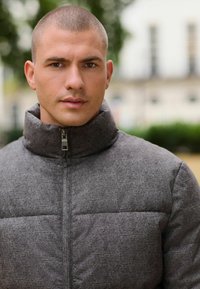 Harmaa pehmustettu takki, jossa on korkea kaulus, vetskari ja teksturoitu kangas, joka esittelee hienovaraisen kuvion. Ihanteellinen viileään säähän.