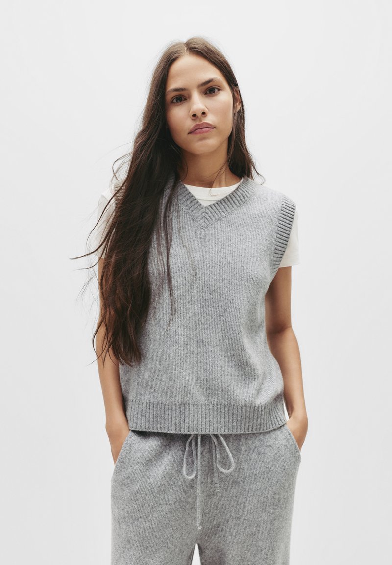 Grauer, gestrickter ärmelloser Pullover mit V-Ausschnitt und gerippten Kanten, kombiniert mit grauen Jerseyhosen mit Kordelzug und einem weichen, strukturierten Finish.