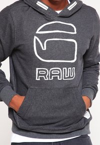 Felpa grigia con tasca frontale, logo bianco "RAW" e cordini a contrasto. Presenta polsini e orlo a coste per una texture aggiuntiva.