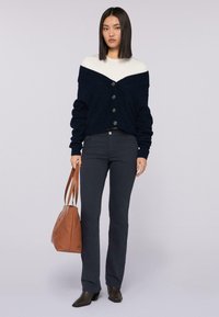 Světlý cardigan v námornické a krémové barvě s pěti knoflíky, spojený s tmavými džíny a hnědou kabelkou. Modelka má na sobě špičaté černé boty.