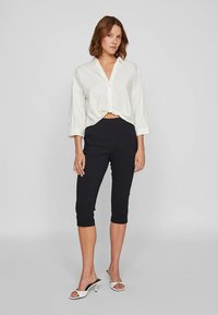 Λευκό πουκάμισο με κουμπιά και δέσιμο μπροστά, συνδυασμένο με μαύρο, κομμένο παντελόνι και λευκά σανδάλια με τακούνι. Λείο ύφασμα, άνετη εφαρμογή.