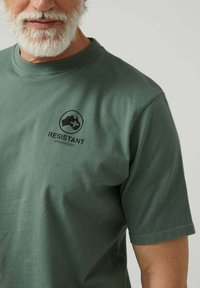 Hombre barbudo que lleva una camiseta verde con el texto y el logotipo "RESISTANT altonadock" en el lado izquierdo del pecho, sobre un fondo simple.