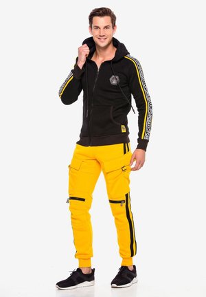 Schwarze Zip-Hoodie mit gelben Streifen und Logo auf der Brust, kombiniert mit leuchtend gelben Cargo-Hosen mit Reißverschlusstaschen und schwarzen Akzenten.