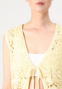 Neulottu keltainen liivi avoimella edustalla ja solmittavalla kiinnityksellä. Sisältää monimutkaisia kuvioita ja tekstuurisen pinnan. Päällä valkoinen paita.