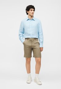 Άνδρας στέκεται με τα χέρια στις τσέπες, φοράει ένα ανοιχτό μπλε πουκάμισο με μακριά μανίκια, μπεζ σορτς, λευκές κάλτσες και λευκά αθλητικά παπούτσια.