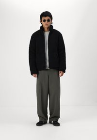 Veste en polaire noire texturée avec col montant, poches avant, portée sur un pull à côtes gris et un pantalon large olive. Chaussures noires.