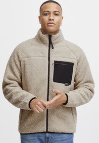 Beige fleece jacket with a high collar, black zippered chest pocket, and black accents. Features textured fabric and contrasting hem detailing.
