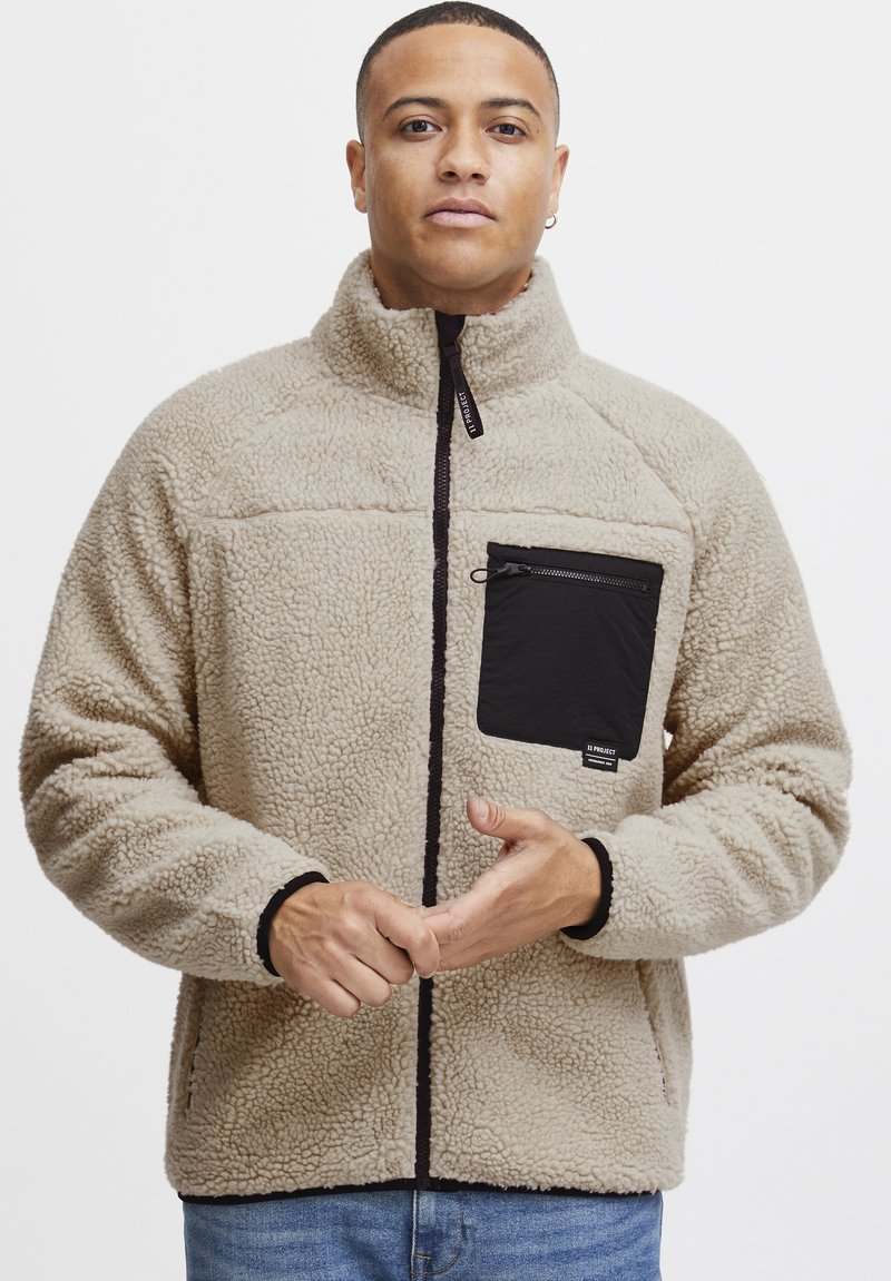 Beige fleece jacket with a high collar, black zippered chest pocket, and black accents. Features textured fabric and contrasting hem detailing.
