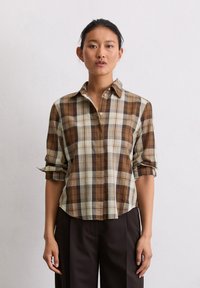 Chemise à carreaux marron et crème avec des manches retroussées, dotée d'un col et d'une coupe décontractée. Associée à un pantalon foncé.