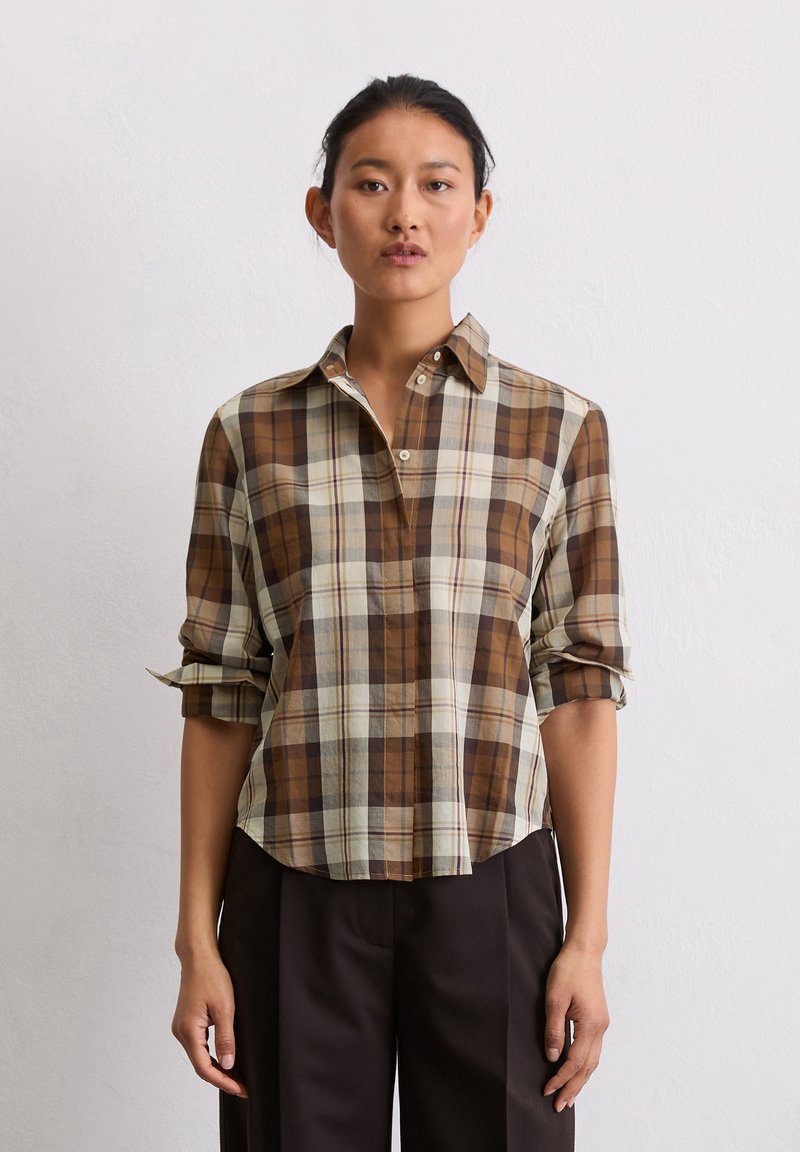 Chemise à carreaux marron et crème avec des manches retroussées, dotée d'un col et d'une coupe décontractée. Associée à un pantalon foncé.