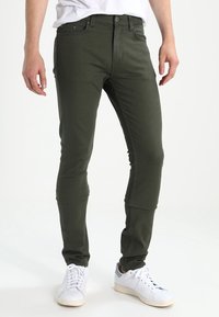 Olijfgroene slim-fit jeans met een gladde textuur, voorzien van voorzakken en een knoopsluiting, gecombineerd met witte sneakers.
