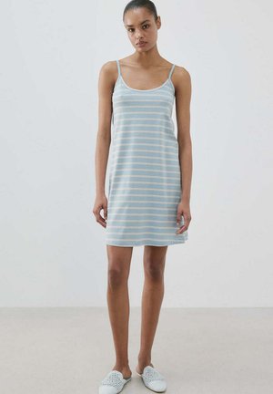 Žena u svijetloplavoj haljini bez rukava s bijelim horizontalnim prugama i bijelim cipelama bez vezica, stoji nasuprot jednostavne pozadine.