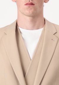Blazer beige con rever a punta, indossato sopra una t-shirt bianca a girocollo. Presenta un sottile dettaglio zip sull'alto del rever del blazer.