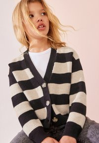 Schwarz-weißer gestreifter Cardigan mit V-Ausschnitt, geripptem Textur und drei Knöpfen an der Vorderseite. Getragen über einem weißen Hemd.