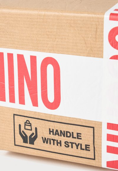 Boîte en carton scellée avec un ruban blanc marqué "INO" en rouge, étiquetée "MANIPULER AVEC STYLE" avec une icône de mains tenant un sac de courses.