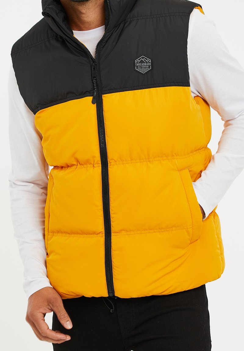 Gilet matelassé avec une moitié supérieure noire et une moitié inférieure jaune vif, doté d'une fermeture éclair à l'avant et d'un logo sur la poitrine.