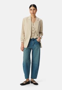 Donna che indossa un blazer beige a righe verticali e un gilet, jeans blu a vita alta, ballerine nere, in piedi contro uno sfondo bianco con una mano in tasca.